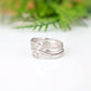 A silver ring with a textured, organic swirl design, resting on a white reflective surface with a soft-focus green background.