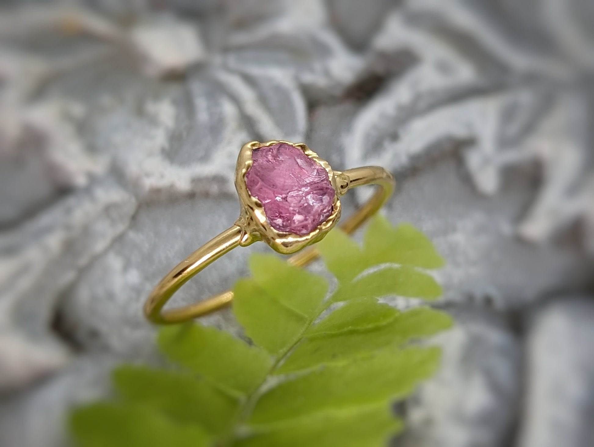 Raw pink spinel ring with golden band, showcasing a natural uncut crystal set against a soft green fern background.