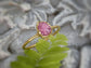 Raw pink spinel ring with golden band, showcasing a natural uncut crystal set against a soft green fern background.