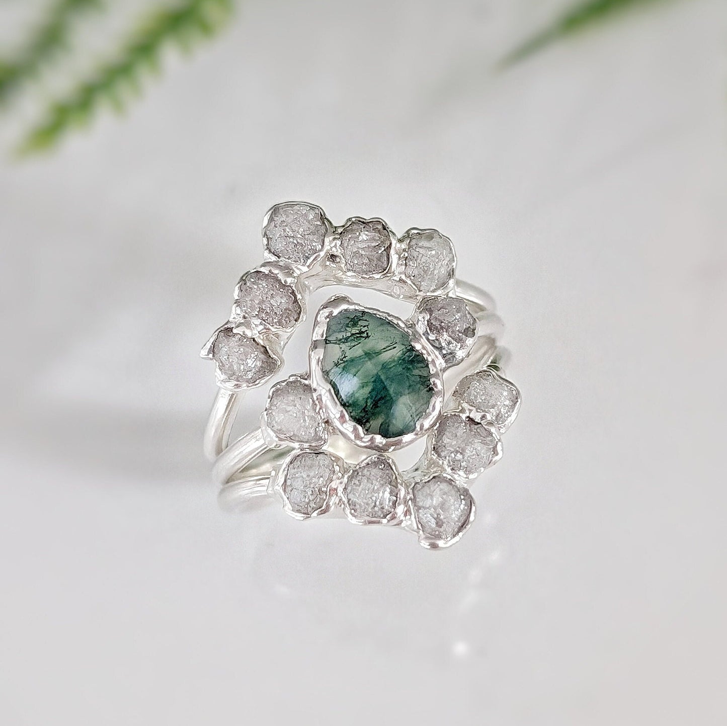 Moss Agate and raw diamond bridal ring set displayed on a reflective surface with greenery backdrop.
