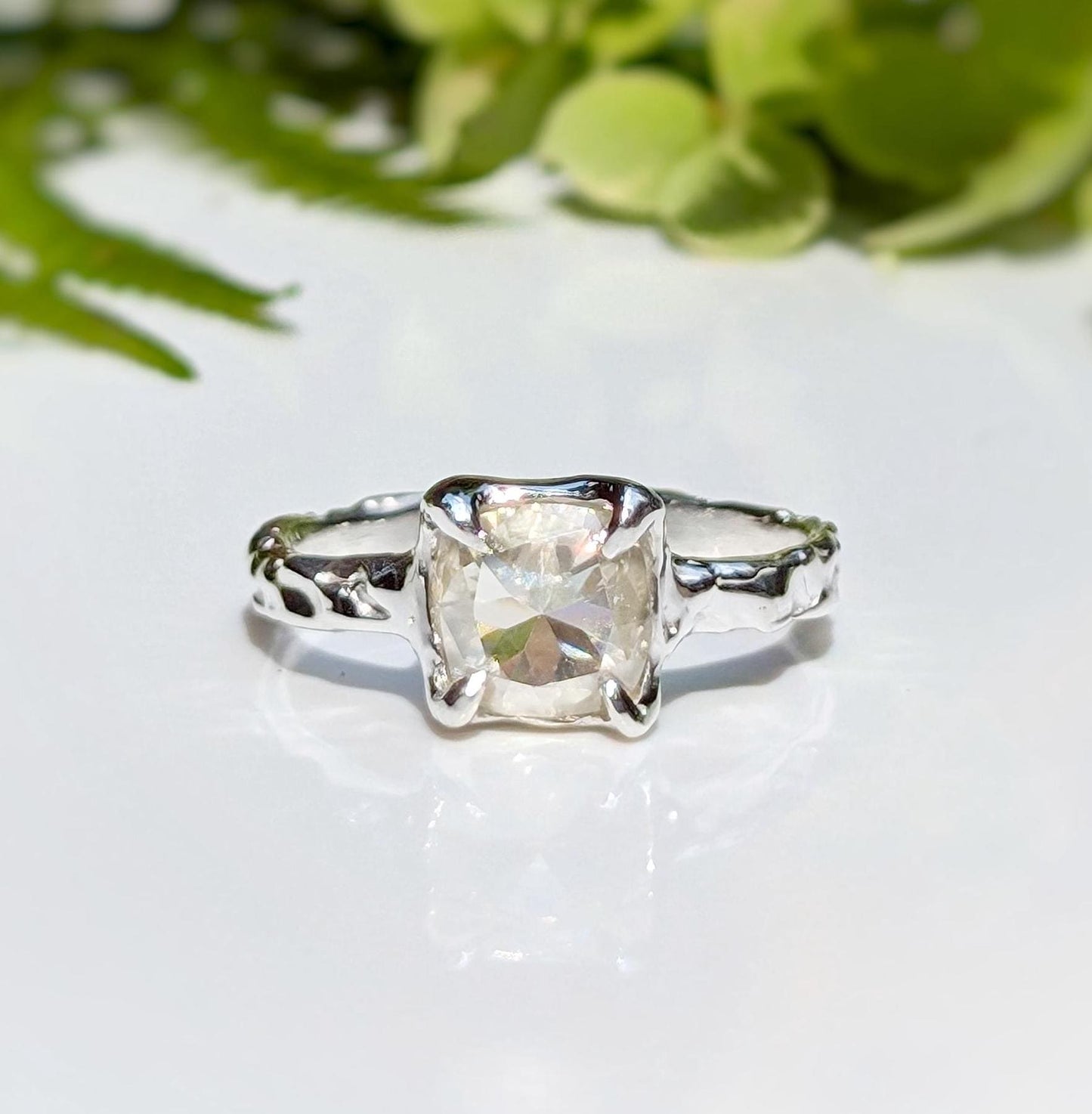 Cushion cut Moissanite engagement ring in textured silver setting with natural greenery in the background.