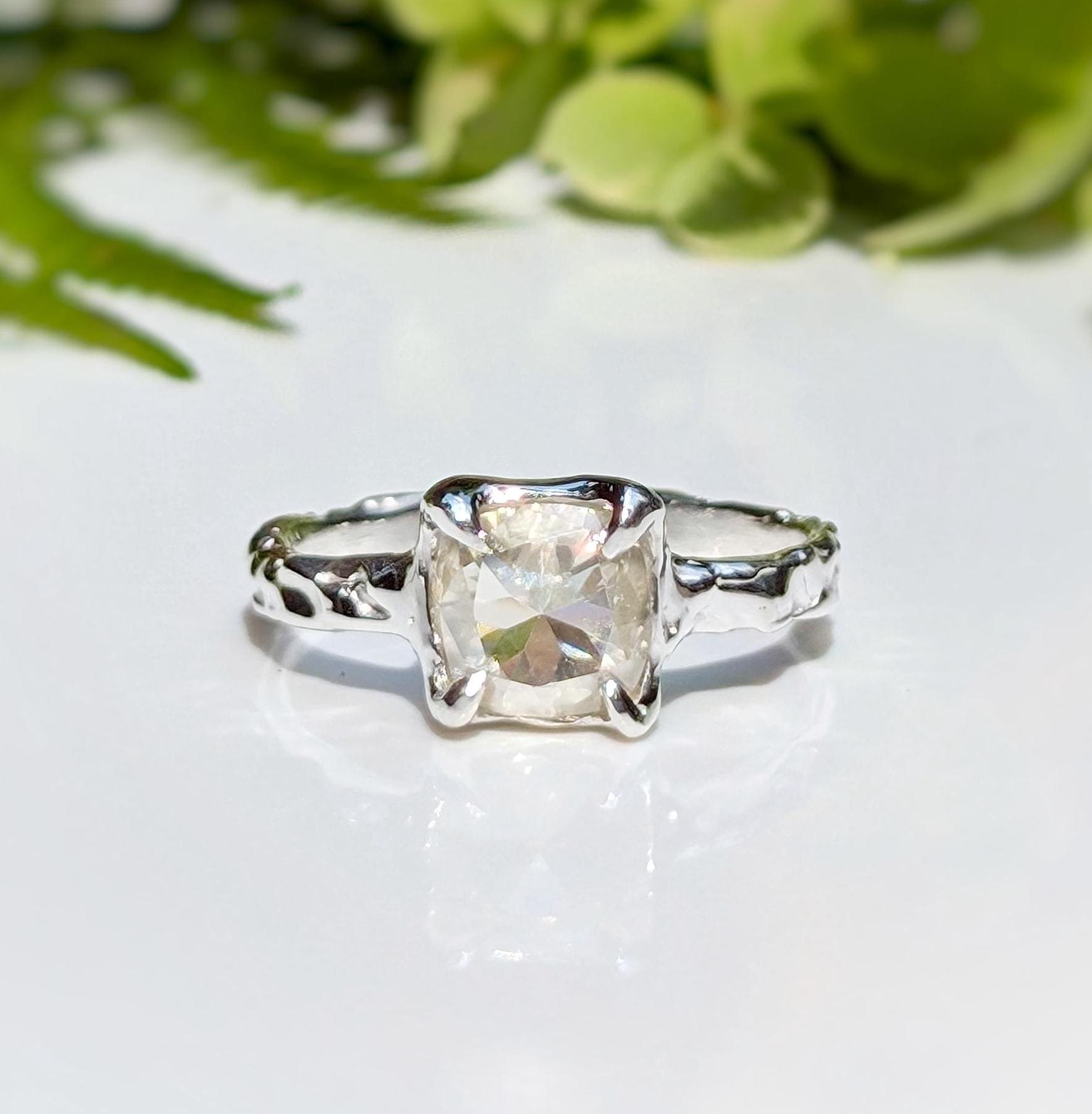 Cushion cut Moissanite engagement ring in textured silver setting with natural greenery in the background.