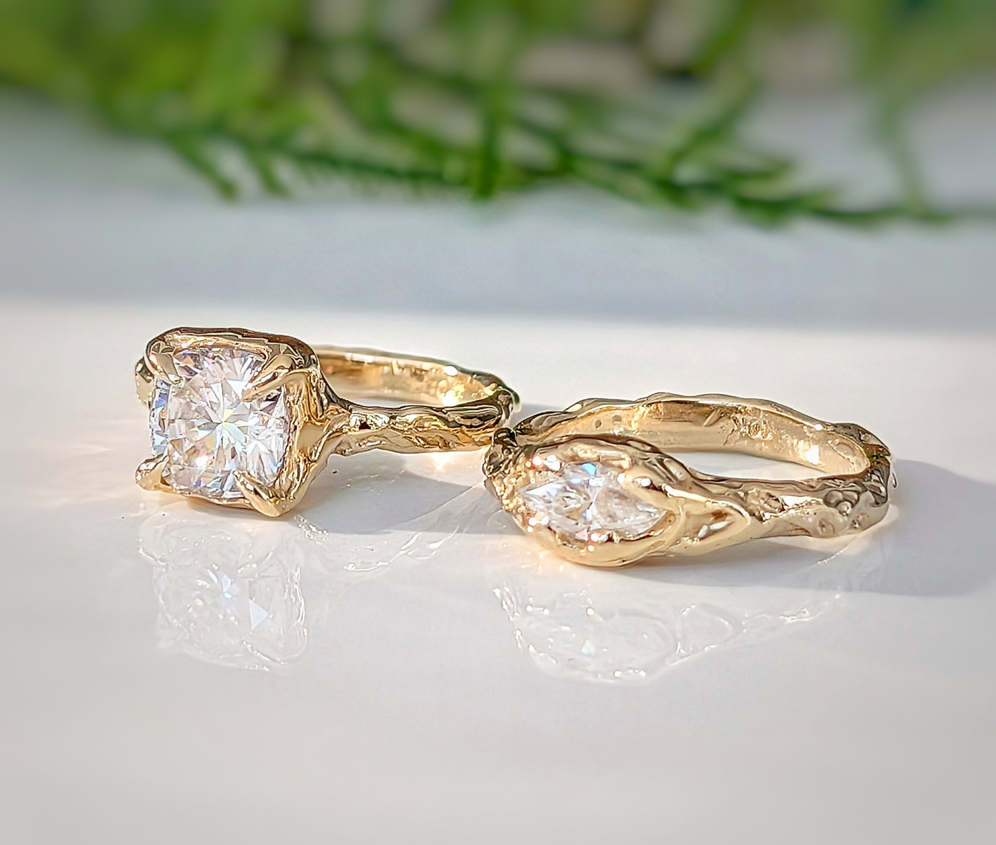 Two gold rings with large, clear gemstones are displayed on a white surface, with a blurred green plant in the background.