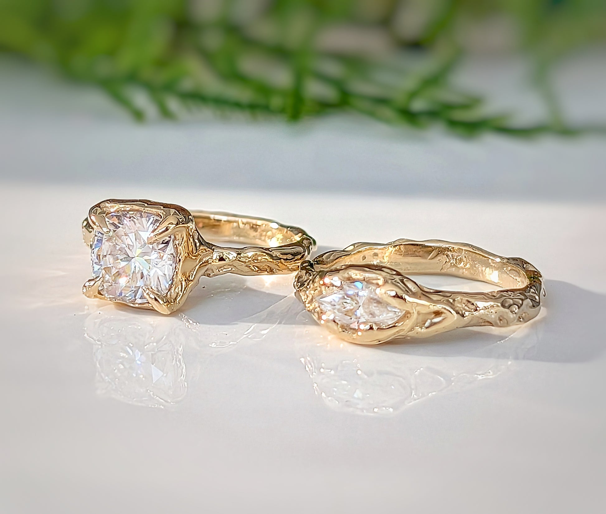 Two gold rings with large, clear gemstones are displayed on a white surface, with a blurred green plant in the background.