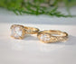 Two gold rings with large, clear gemstones are displayed on a white surface, with a blurred green plant in the background.