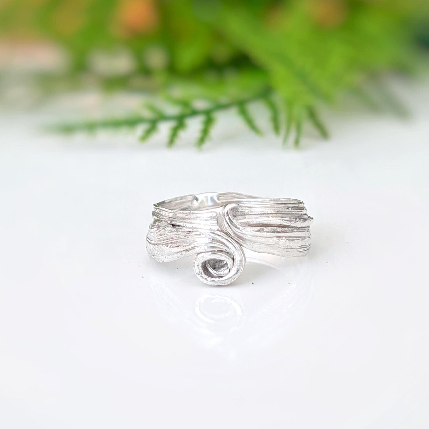 A silver ring with a textured, organic swirl design, resting on a white reflective surface with a soft-focus green background.