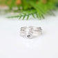 A silver ring with a textured, organic swirl design, resting on a white reflective surface with a soft-focus green background.