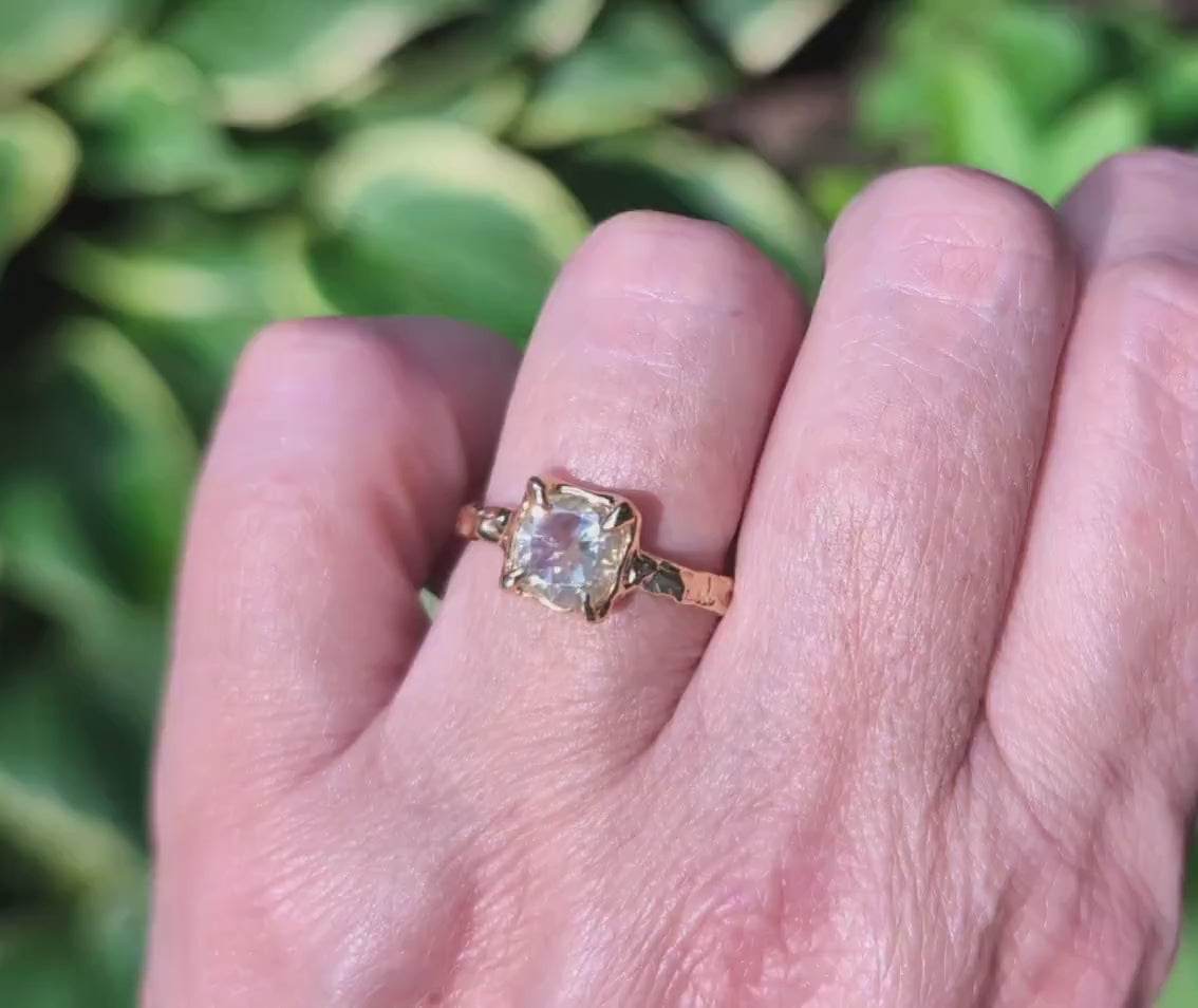 Cushion cut Moissanite engagement ring in textured gold setting worn on hand.