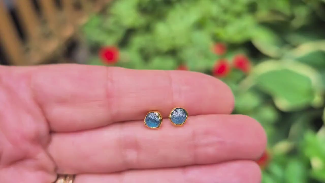 A hand holding a pair of natural teal blue Montana Sapphire stud earrings - set in an outdoor setting with greenery in the background