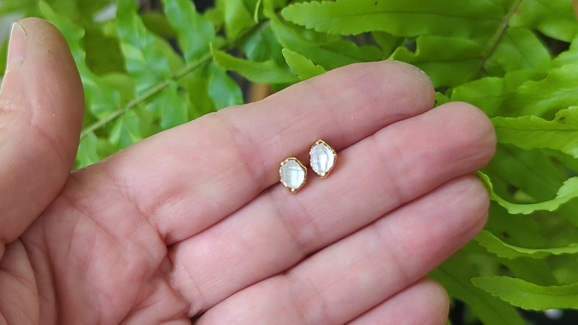 Handcrafted Herkimer diamond stud earrings held in hand, surrounded by green ferns, showcasing their unique design.