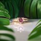 Rubellite Tourmaline engagement ring in solid 14k gold, featuring a trilliant cut and textured band, placed among lush green leaves.