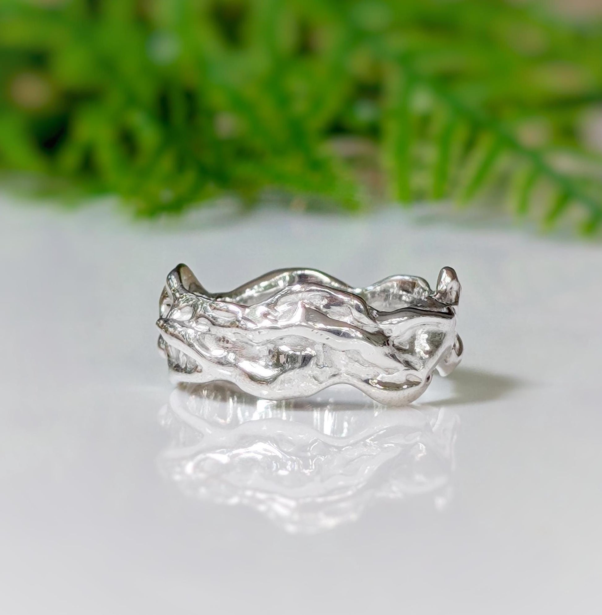 Handcrafted sterling silver molten textured wedding band on a reflective surface with greenery in the background.