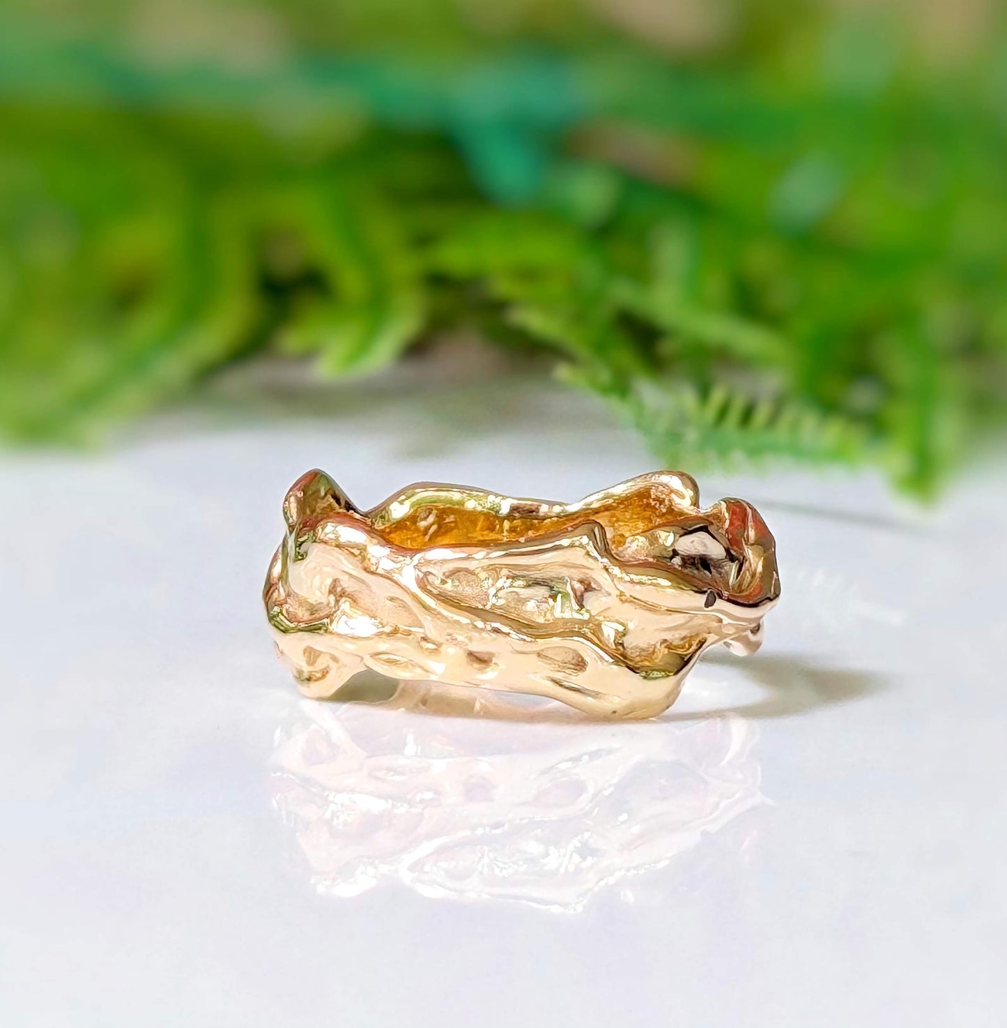 Textured molten gold wedding band resting on a reflective surface with greenery in the background.