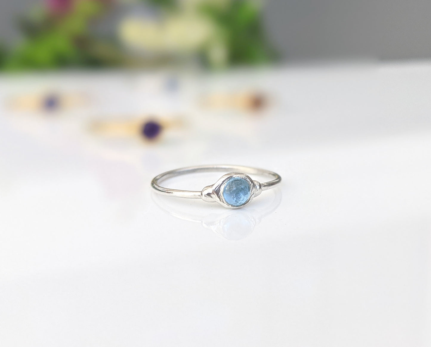 Dainty blue Topaz stacking ring on a reflective surface, surrounded by other minimalist rings.