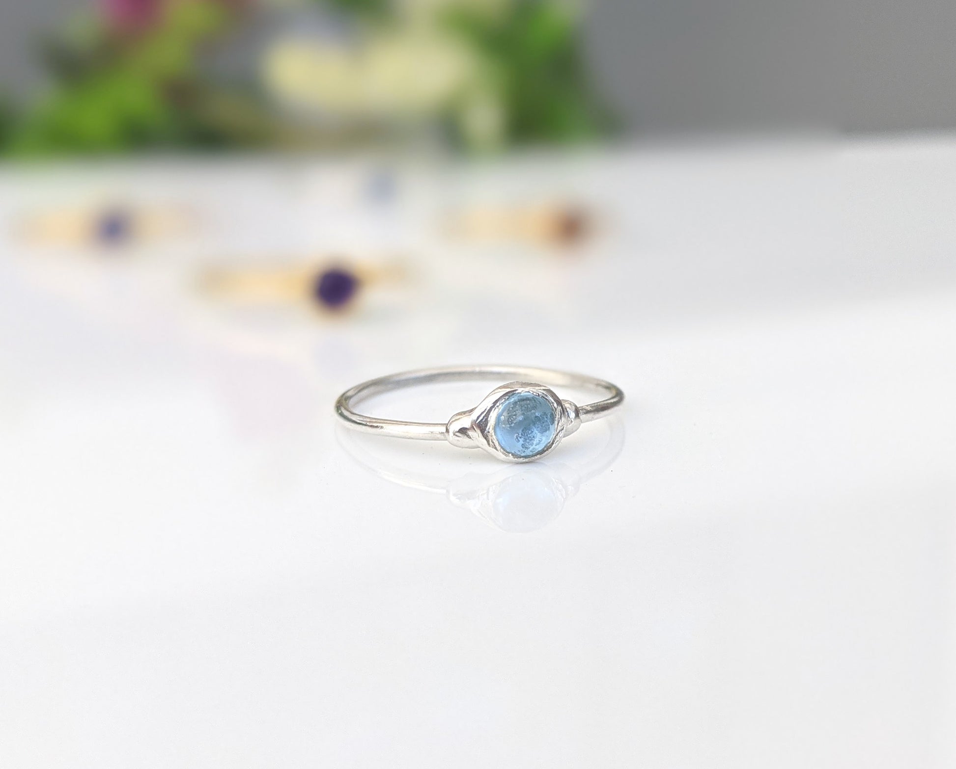 Dainty blue Topaz stacking ring on a reflective surface, surrounded by other minimalist rings.