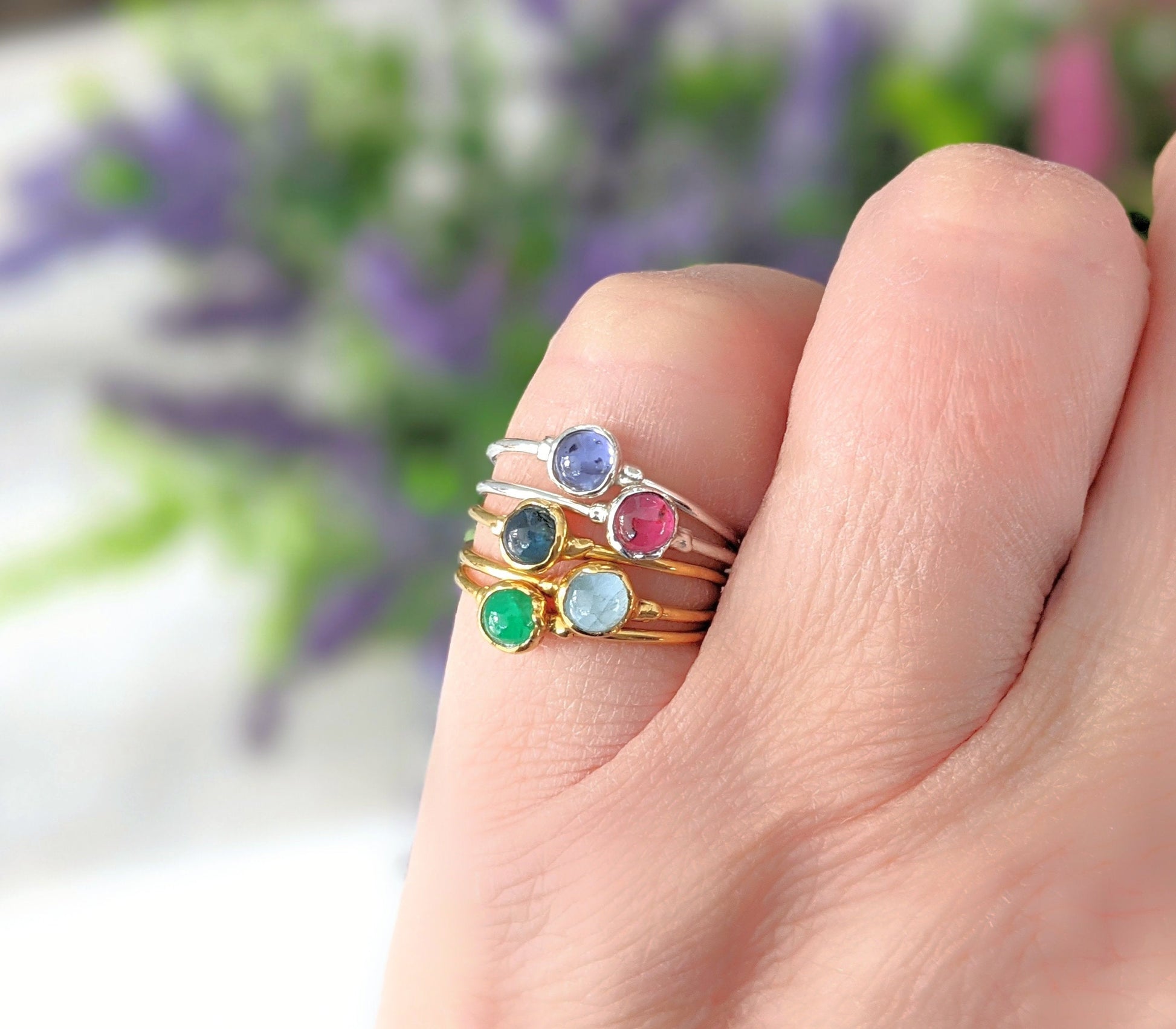 A hand displaying multiple dainty stacking rings featuring colorful gemstones including Sapphire, Ruby, and Emerald.