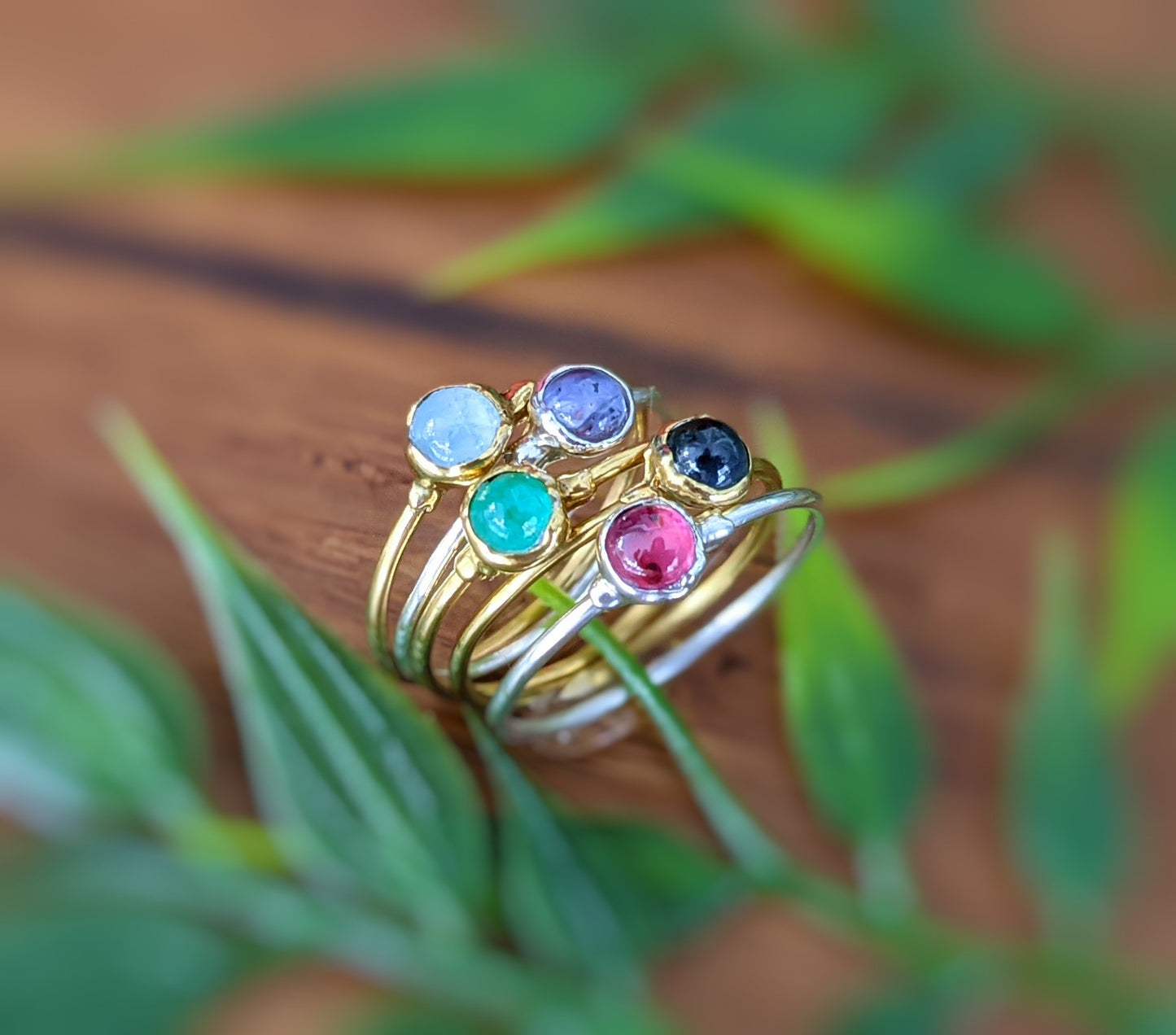 Stack of colorful gemstone rings featuring blue, green, black, and pink stones on a wooden surface with greenery.