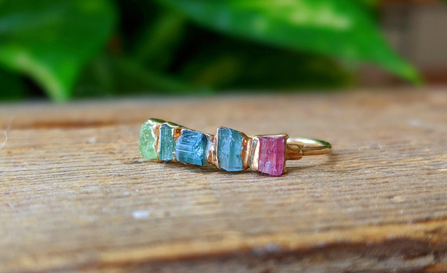 Colorful raw Tourmaline eternity ring on wood background, featuring green, blue, and pink stones set in a gold band.