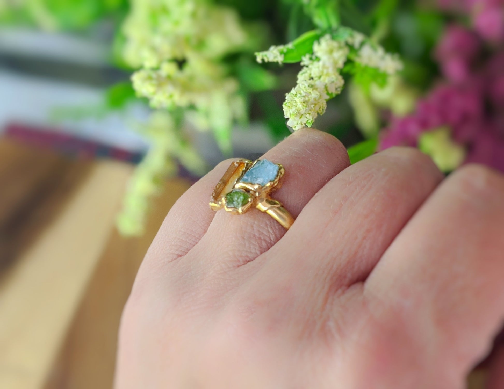 Raw Topaz, Aquamarine, and Peridot cluster ring on a hand, adorned with fresh flowers in the background.