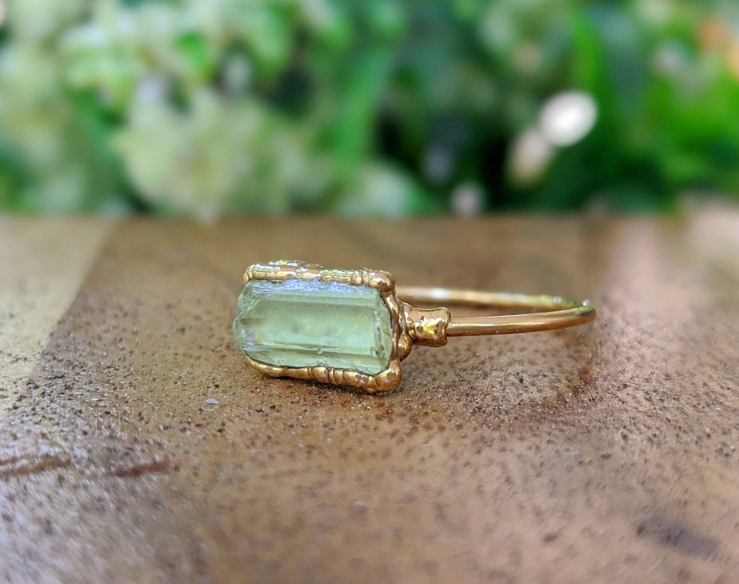 Raw yellow Apatite ring on a gold band, highlighting natural beauty, set on a wooden surface with greenery in background.