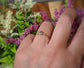 Hand wearing a handcrafted raw blue diamond engagement ring on a gold band, surrounded by flowers.