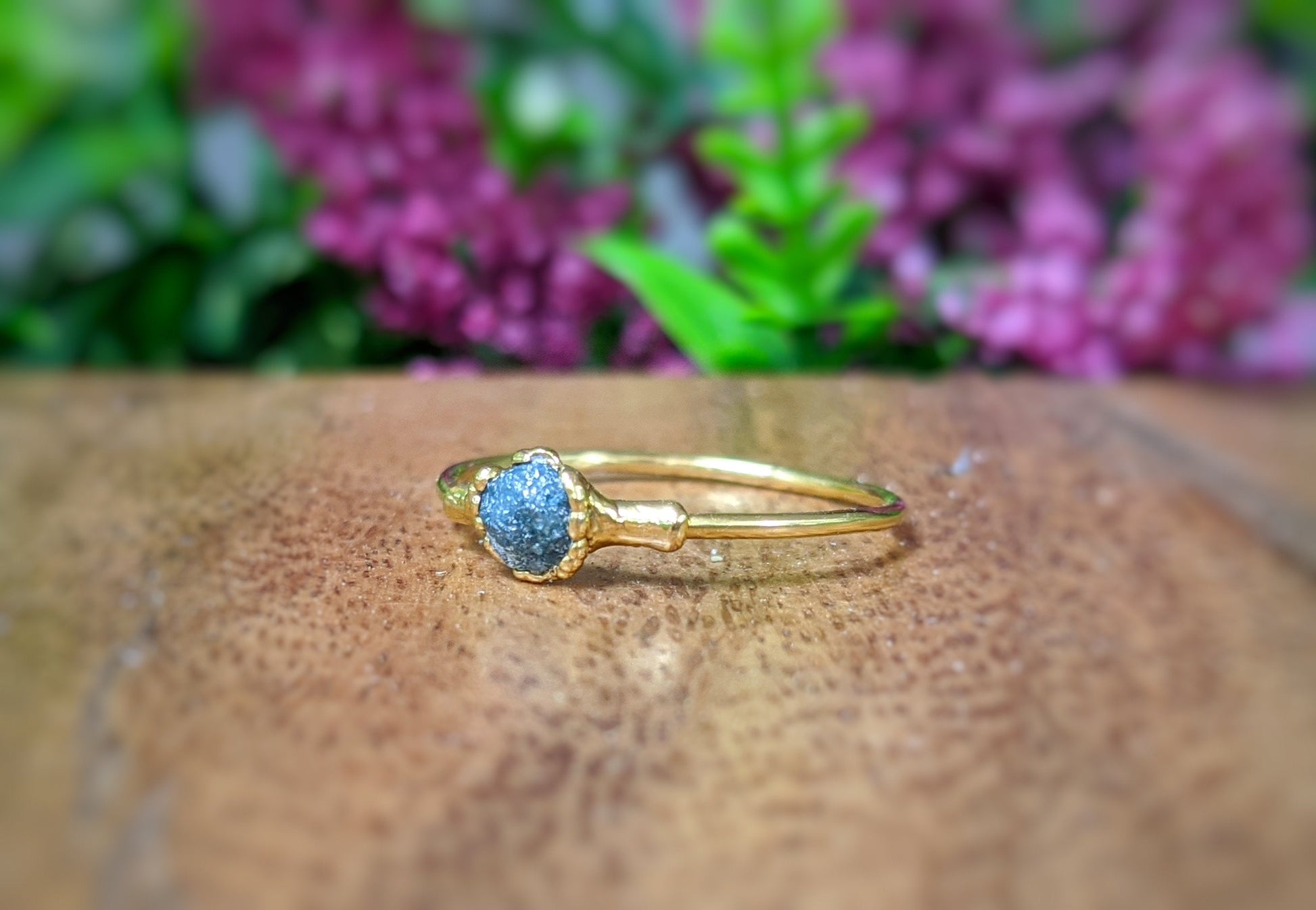 Handcrafted raw blue diamond engagement ring on 18k gold band, displayed against a floral background.