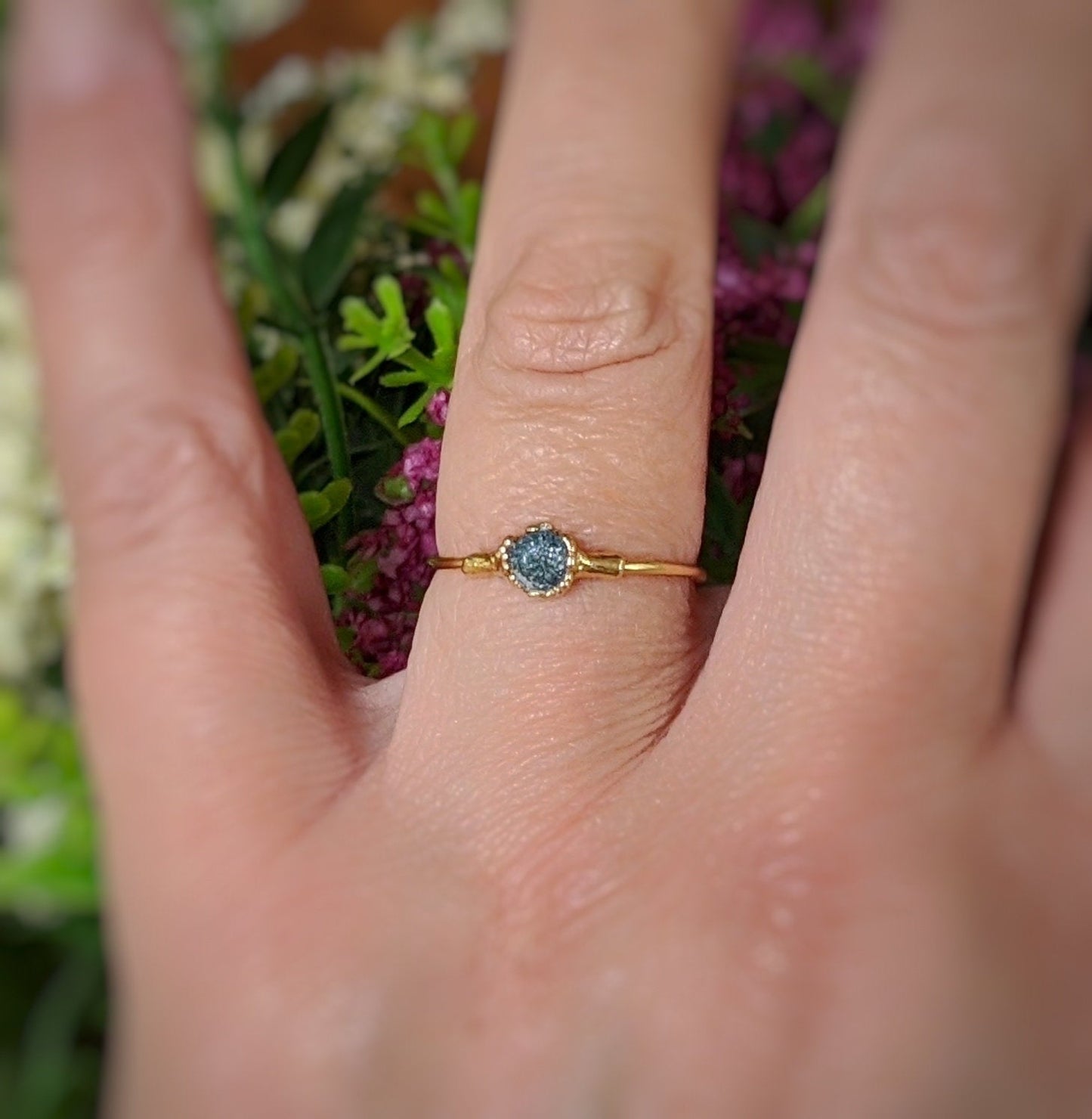 Raw blue diamond engagement ring on a hand, featuring a solid 18k gold band and surrounded by flowers.