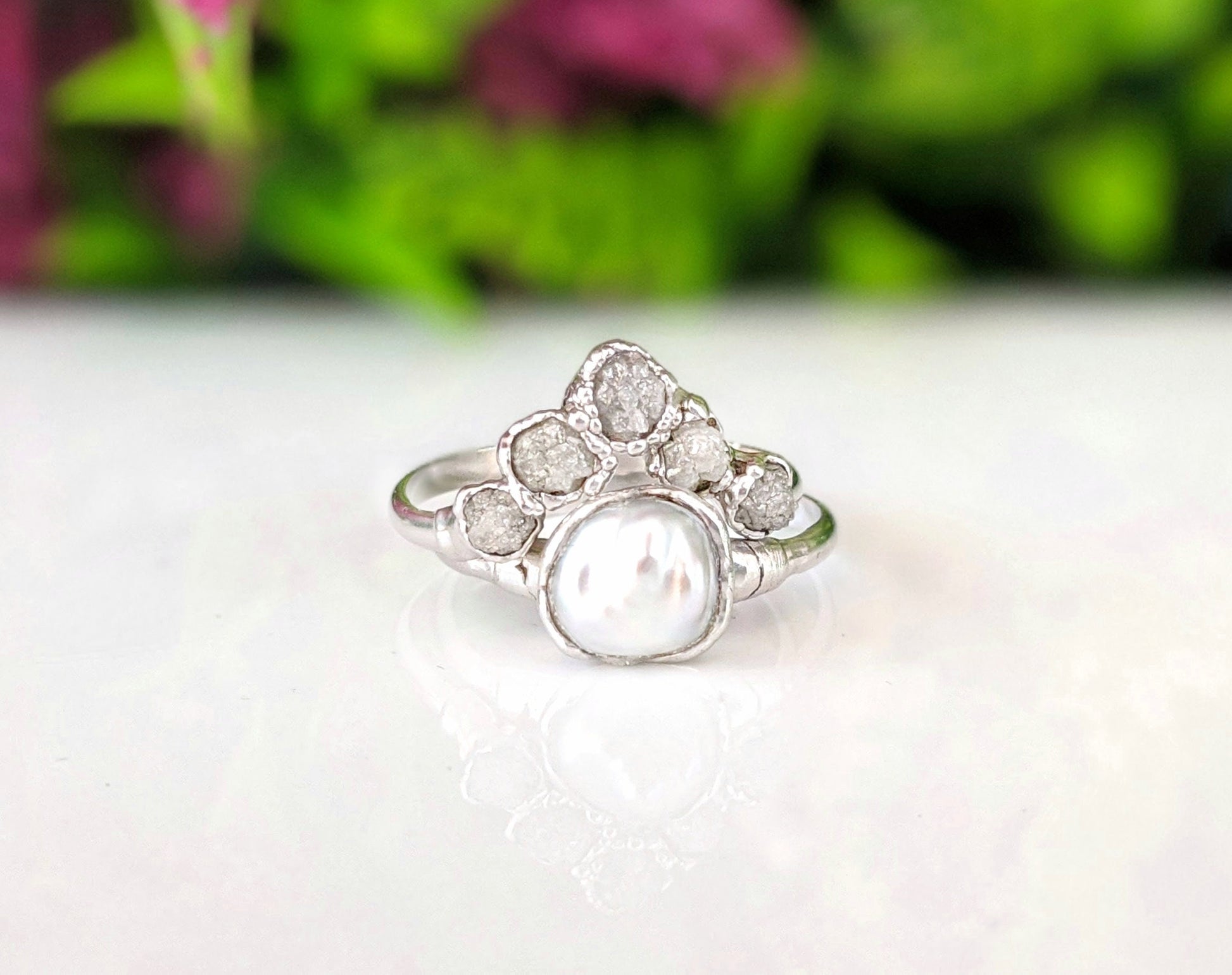 Handcrafted raw diamond and Keshi pearl Chevron wedding ring set on a reflective surface with greenery in the background.