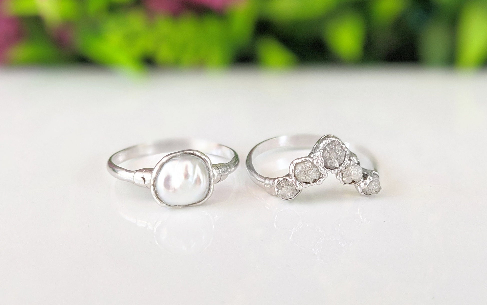 Raw diamond and Keshi pearl Chevron wedding ring set on a reflective surface with greenery in the background.