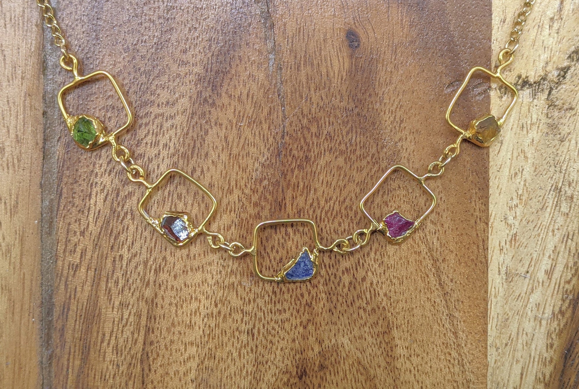 Unique gold square pendant necklace featuring colorful raw gemstones on a wooden background.