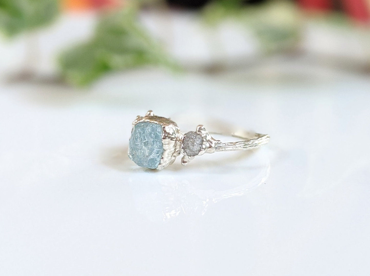 Raw aquamarine and diamond twig ring on a glossy surface with natural greenery in the background.