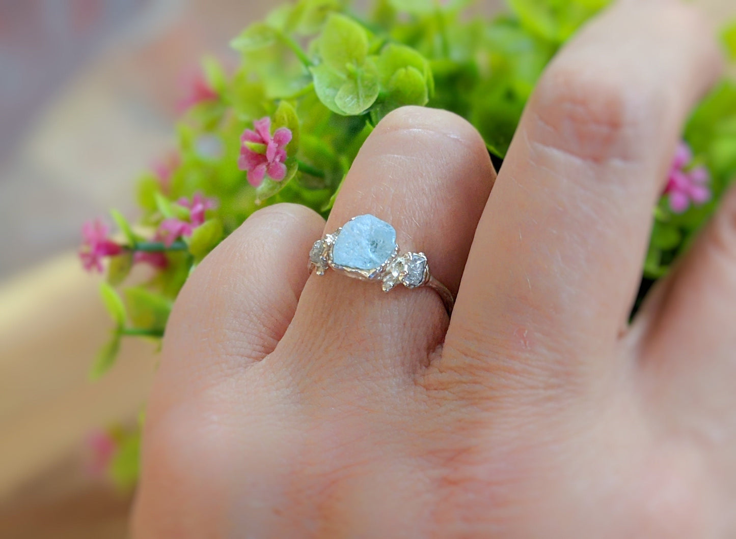 Hand wearing a unique raw aquamarine and diamond twig ring surrounded by green foliage and flowers.