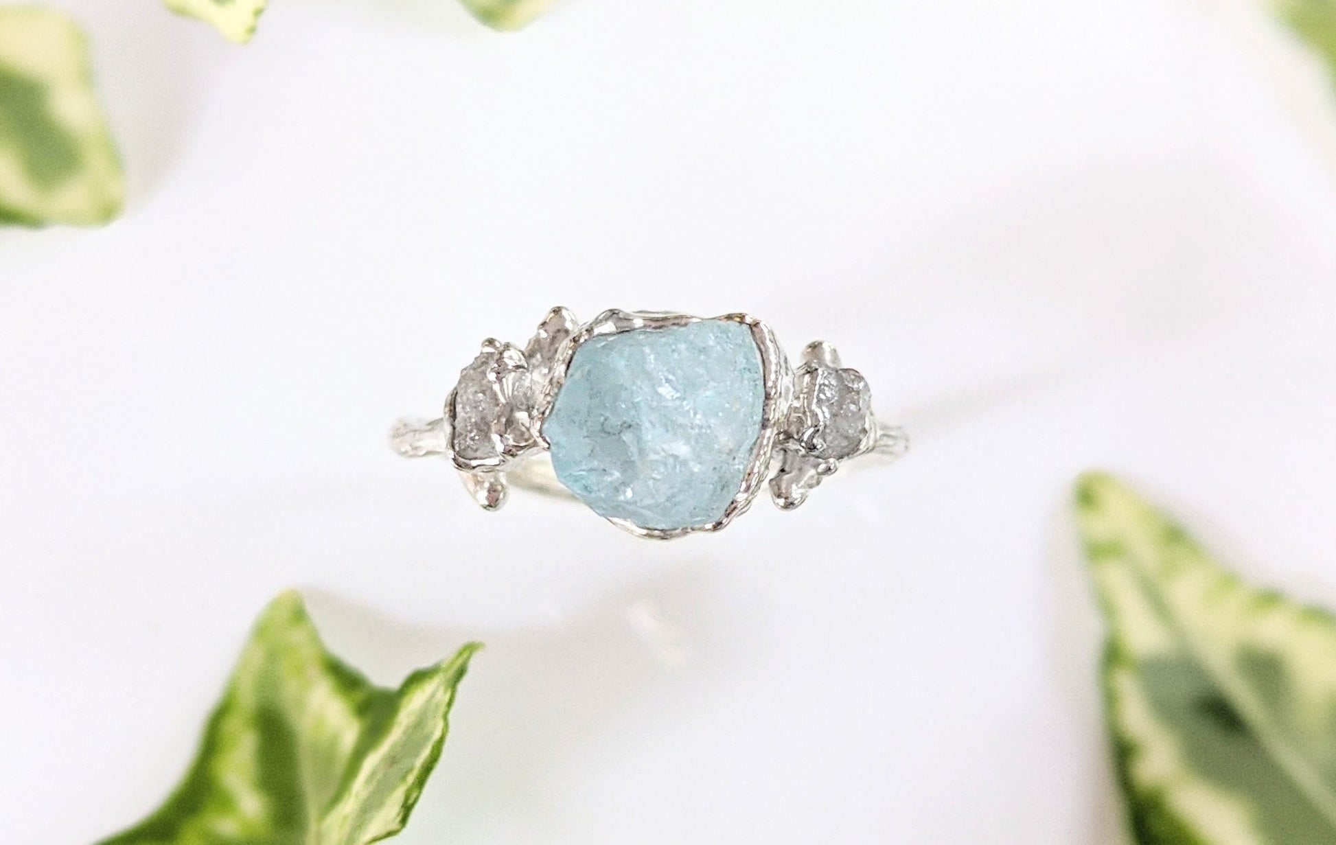 Raw Aquamarine and diamond twig ring set on a Sterling Silver band, surrounded by green leaves.