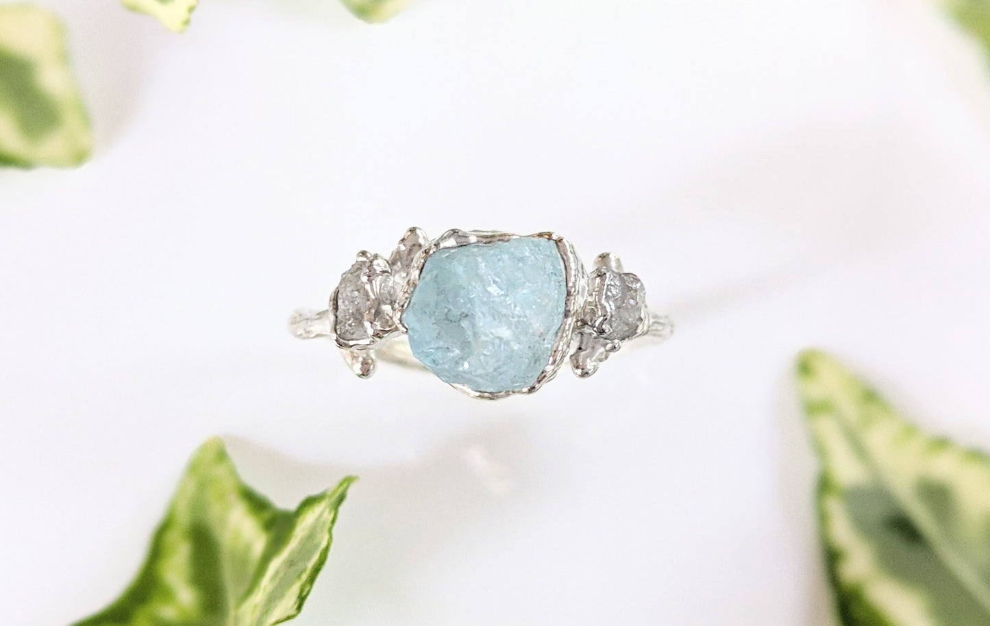 Raw aquamarine and diamond twig ring on a white surface, surrounded by green leaves, featuring a unique design.