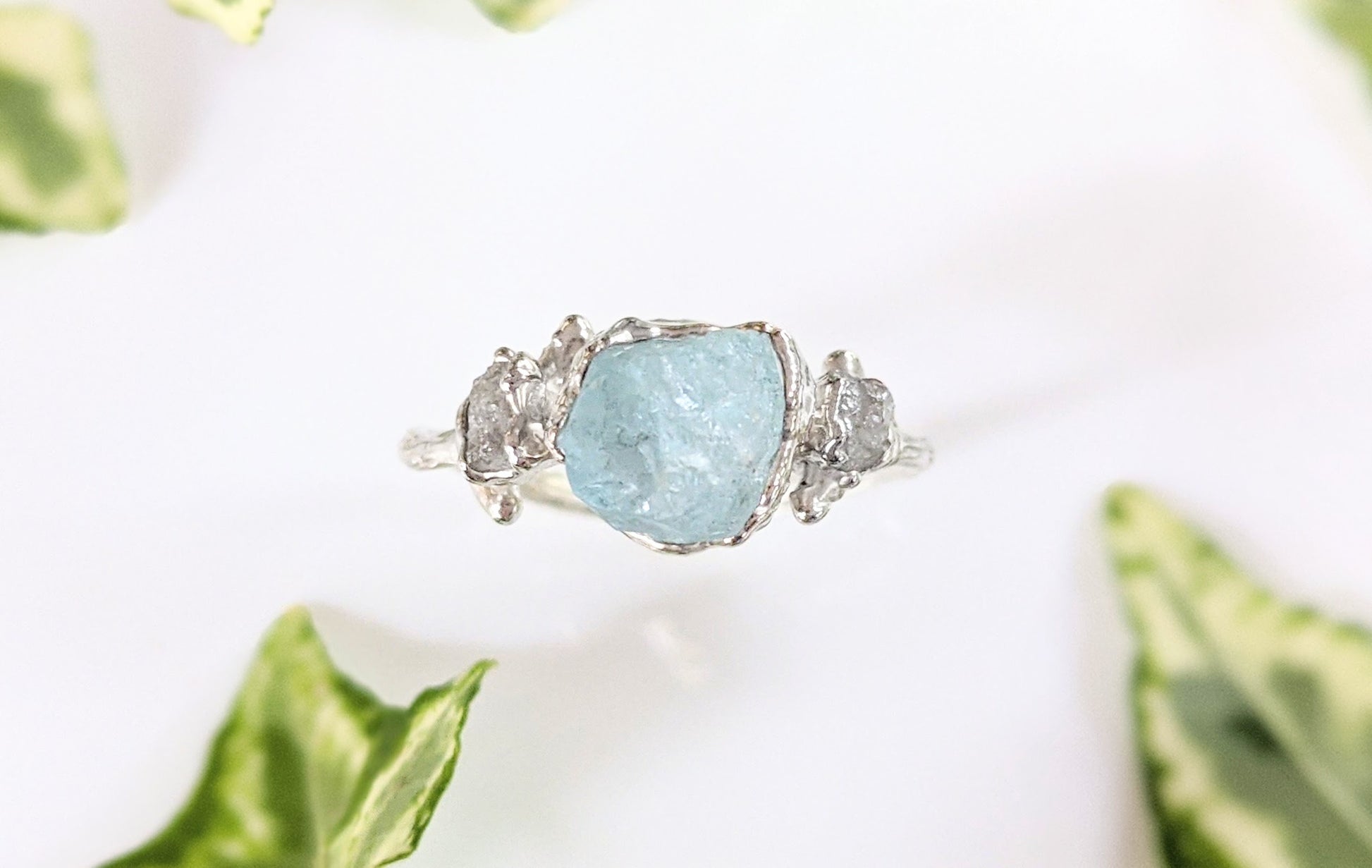 Raw aquamarine and diamond twig ring on a white surface, surrounded by green leaves, featuring a unique design.