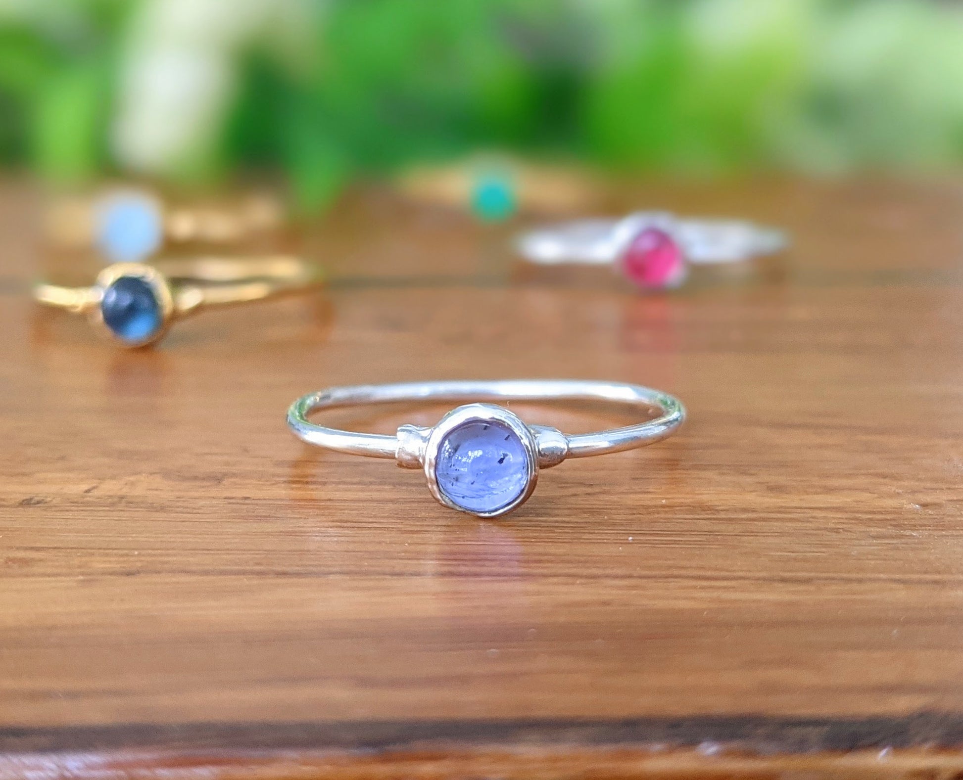 Dainty Tanzanite stacking ring on wooden surface, featuring a natural purple-blue gemstone surrounded by other colorful rings.