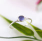 Dainty Tanzanite stacking ring on a green leaf, featuring a small round natural purple/blue gemstone in a silver band.