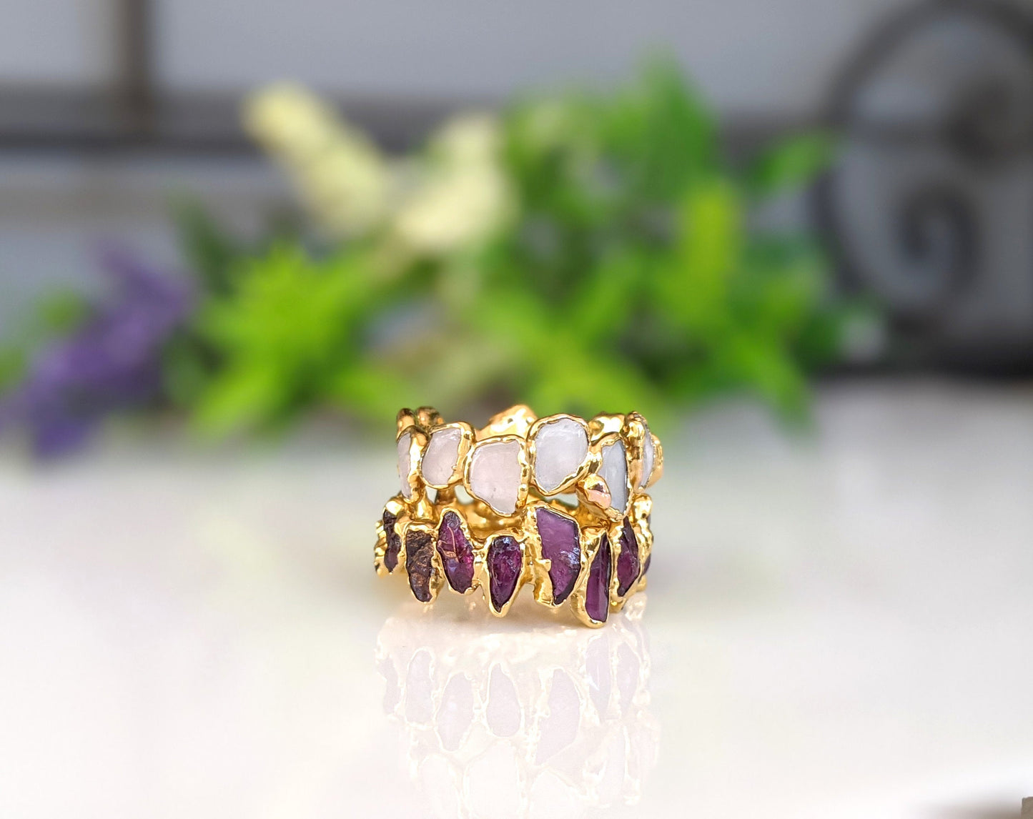 Raw Rhodolite Garnet Eternity Ring with gold filled band and colorful gemstones on a blurred floral background.