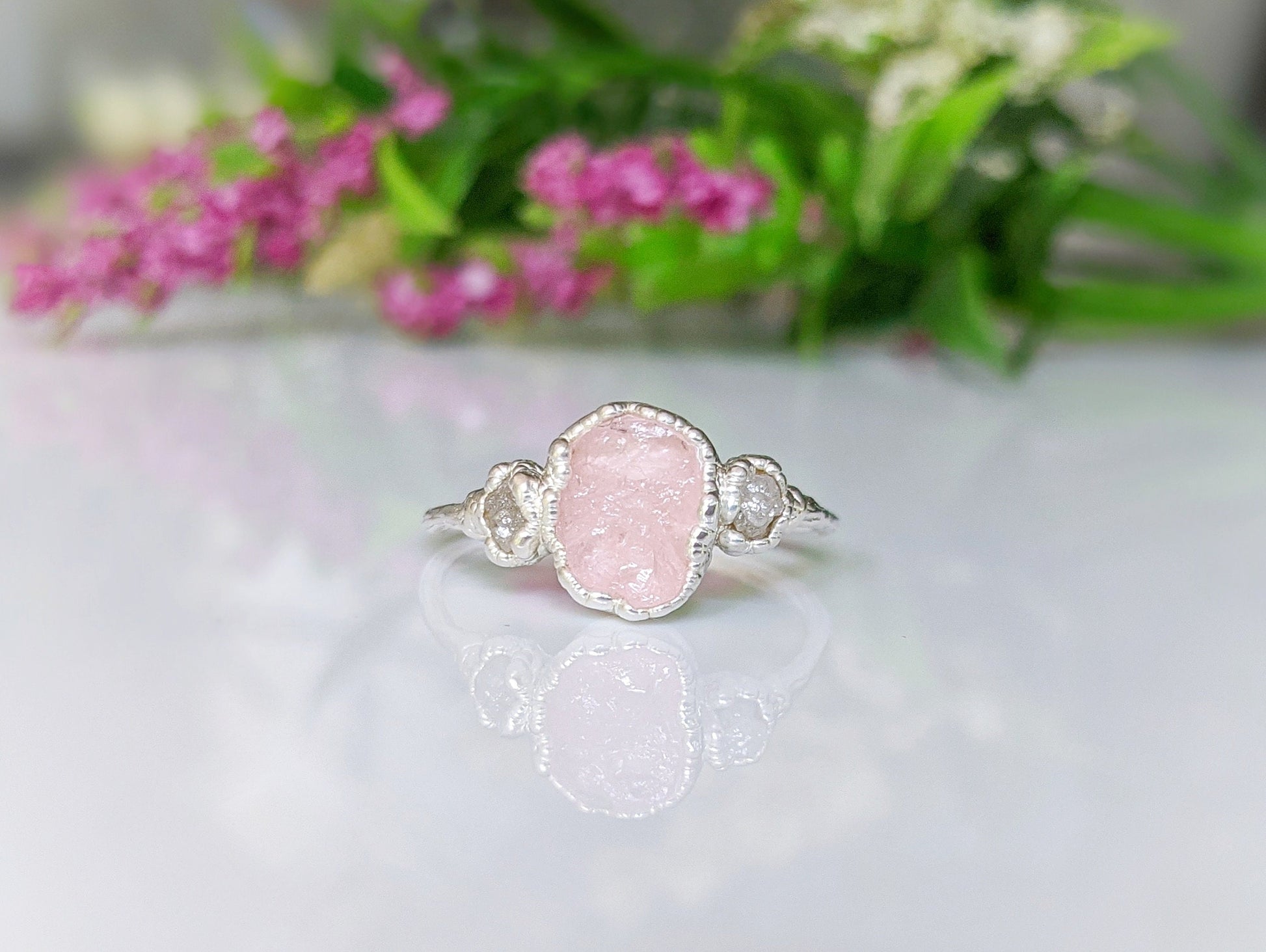 Raw Morganite ring with rough diamond and pink morganite on sterling silver band, set against floral background.