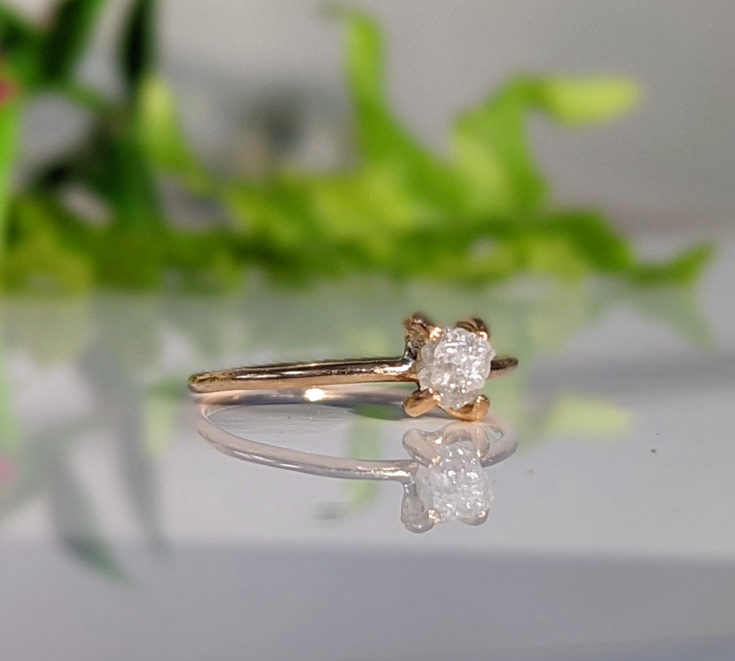 Delicate raw diamond engagement ring on a reflective surface, featuring a 14k gold band and sparkling white diamond.