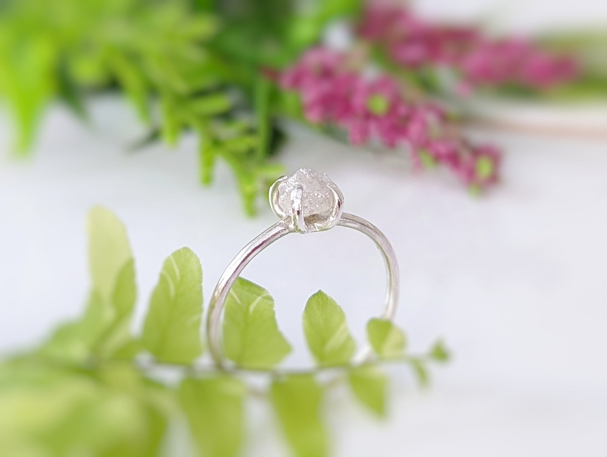 Elegant raw diamond engagement ring with a silver band, surrounded by greenery and soft floral elements.