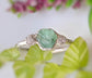 Handcrafted raw emerald and rough diamond engagement ring with rustic charm and elegance, surrounded by greenery.