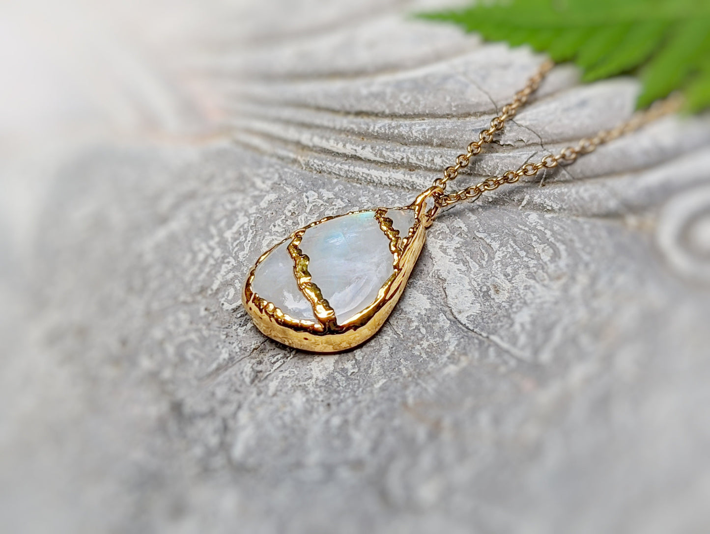 Kintsugi necklace with pear-shaped rainbow Moonstone and 18k gold plating, resting on a textured surface.