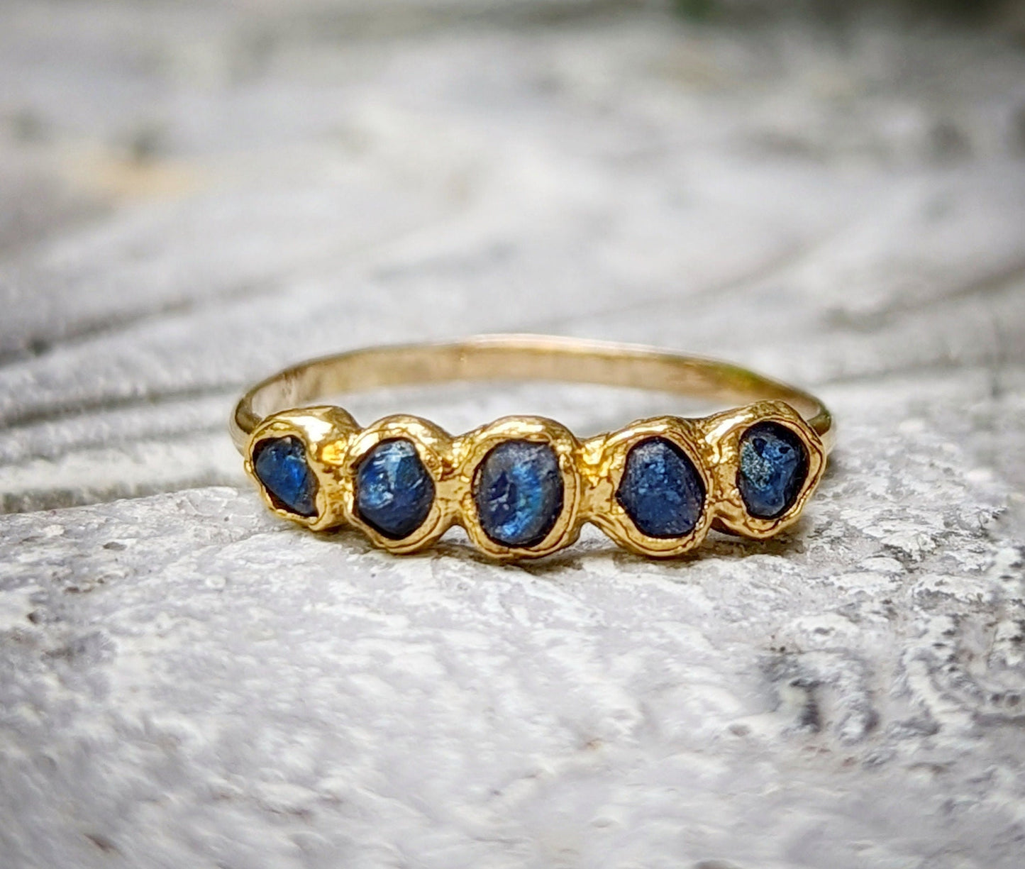 Raw sapphire multi-stone eternity ring on a textured surface, featuring small blue sapphires in a 14k gold filled band.