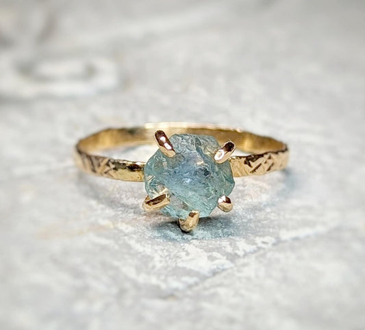 Raw Montana Sapphire engagement ring with textured gold band, showcasing a unique teal-blue gemstone.