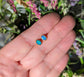 Blue Australian Opal stud earrings in 18k Gold, held in hand against a backdrop of greenery.