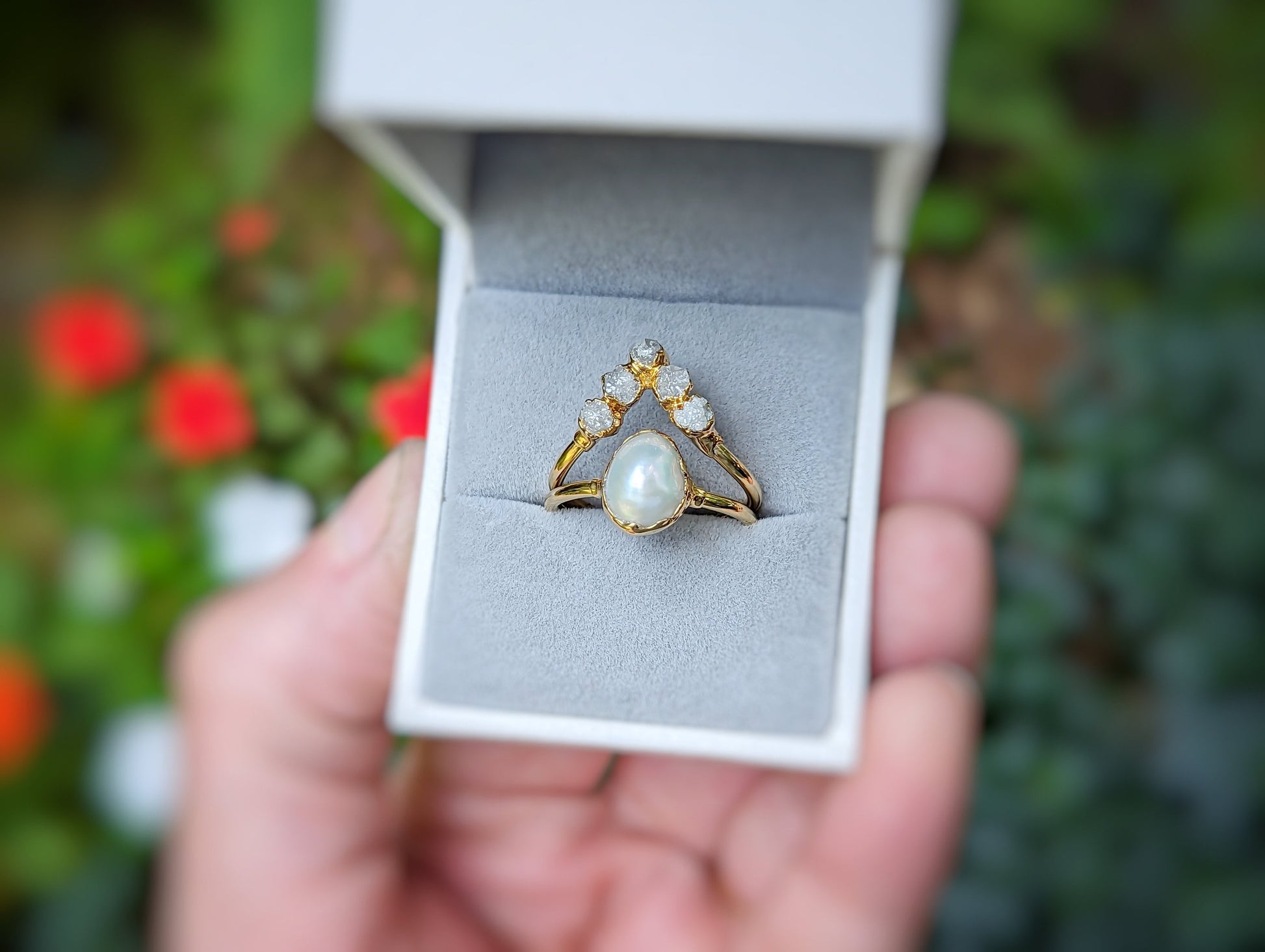 Freshwater pearl and raw diamond chevron wedding ring set displayed in a ring box with vibrant flowers in the background.