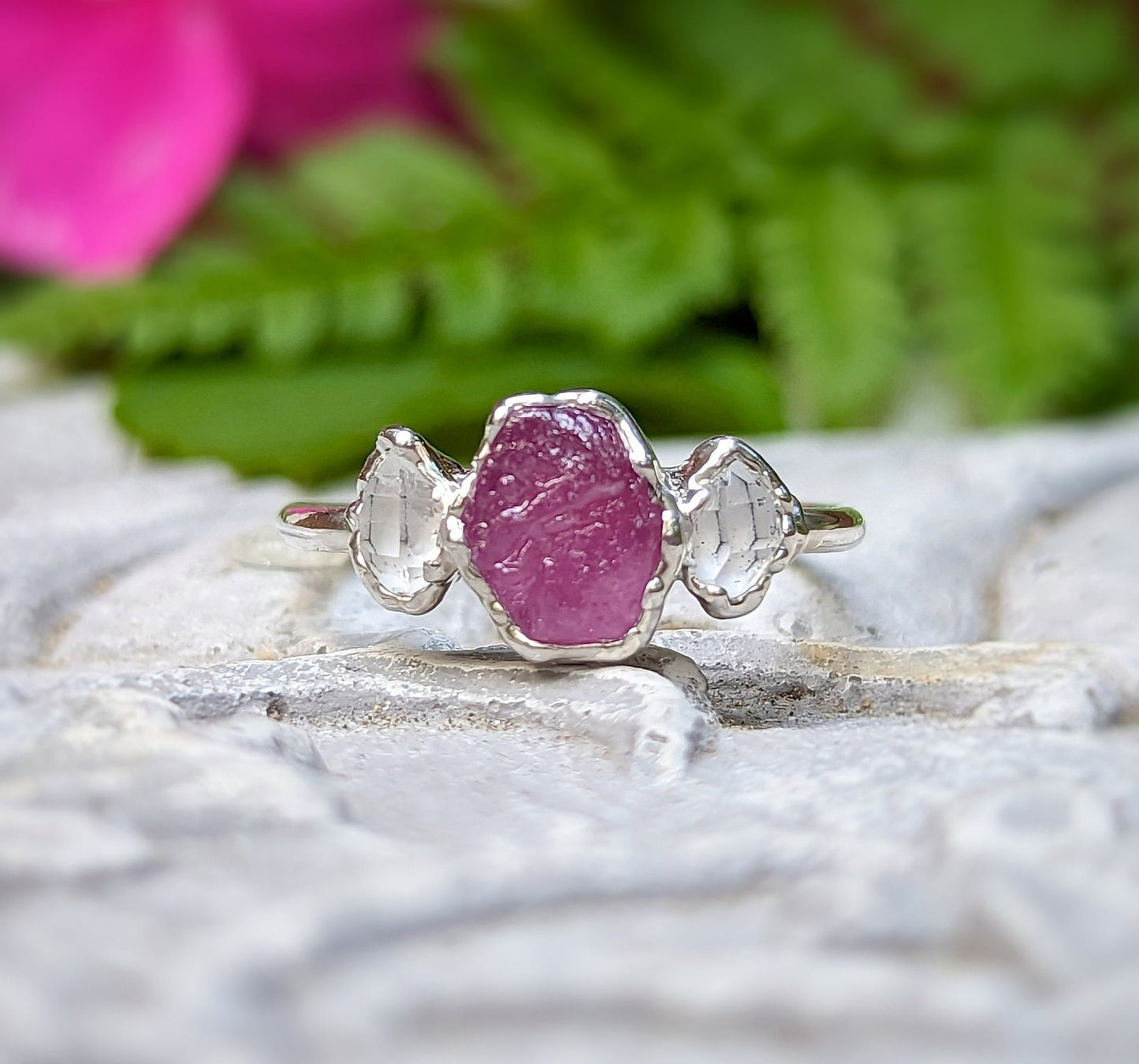 Raw pink Sapphire and Herkimer diamond engagement ring on a stone surface with greenery in the background.