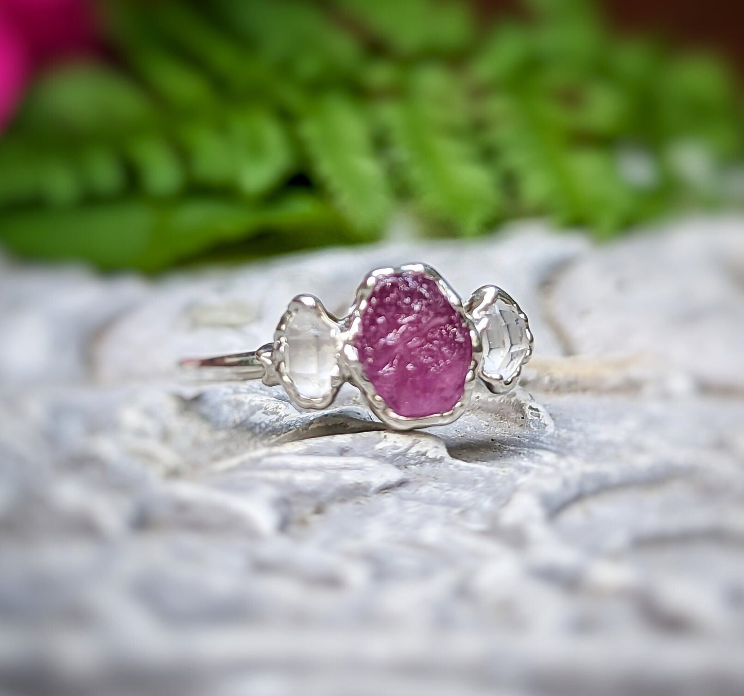 Handcrafted raw pink Sapphire and Herkimer diamond engagement ring on a textured surface surrounded by greenery.