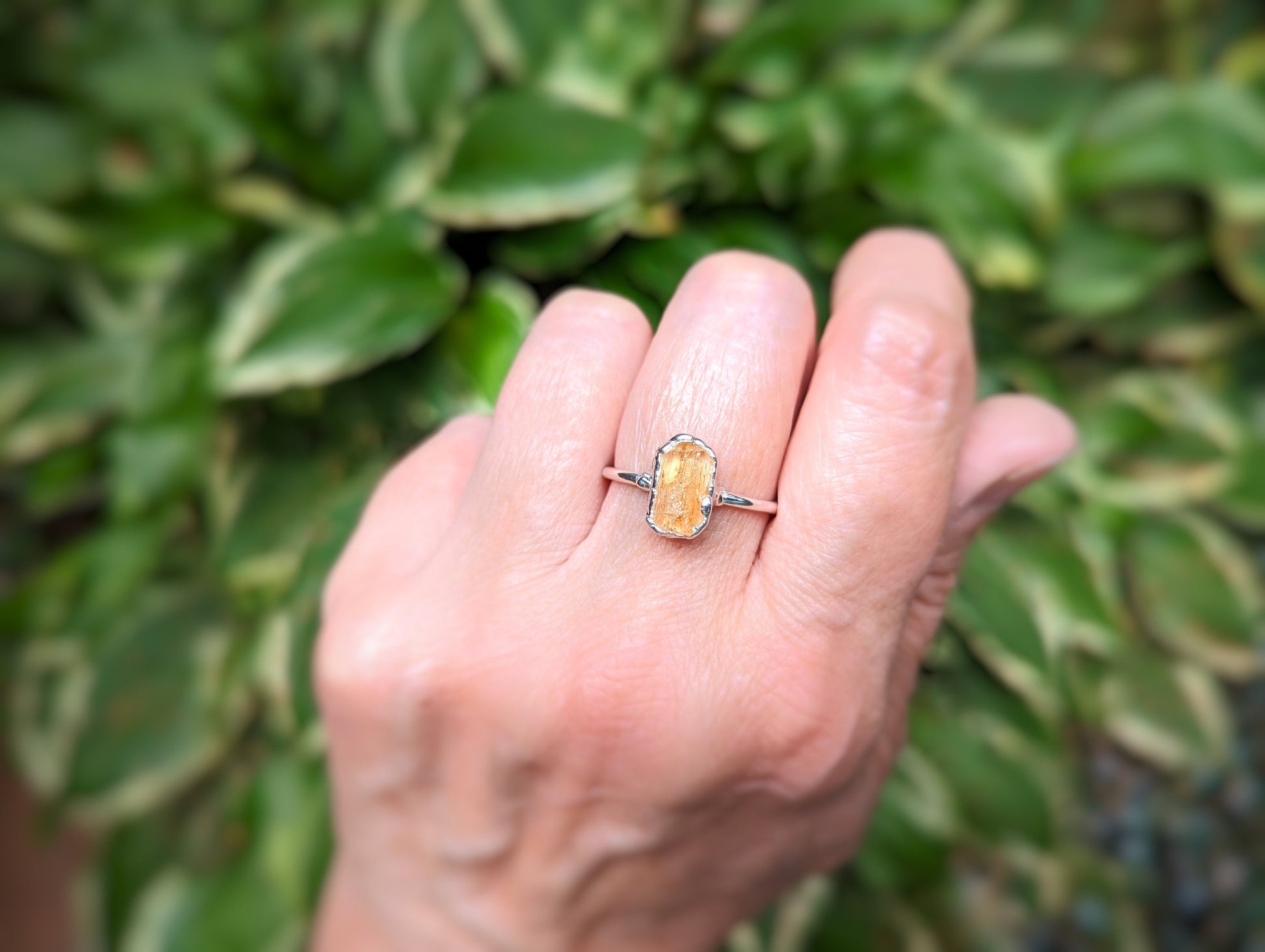 Hand wearing a raw Golden Imperial Topaz ring on a handcrafted Sterling Silver band, amidst green foliage.